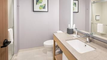 Un baño moderno con cabina de ducha de vidrio, inodoro blanco, lavabo con encimera de mármol y espejo con una obra de arte enmarcada.
