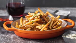 Un plato de papas fritas crujientes en una olla roja, con un recipiente pequeño de salsa y una copa de vino tinto al fondo.