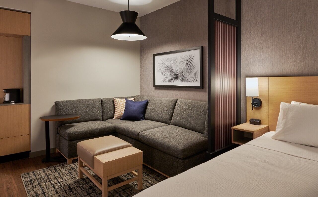 A modern hotel room with a sectional sofa, a small table, a bedside table with a lamp, a bed, and a framed artwork on the wall.