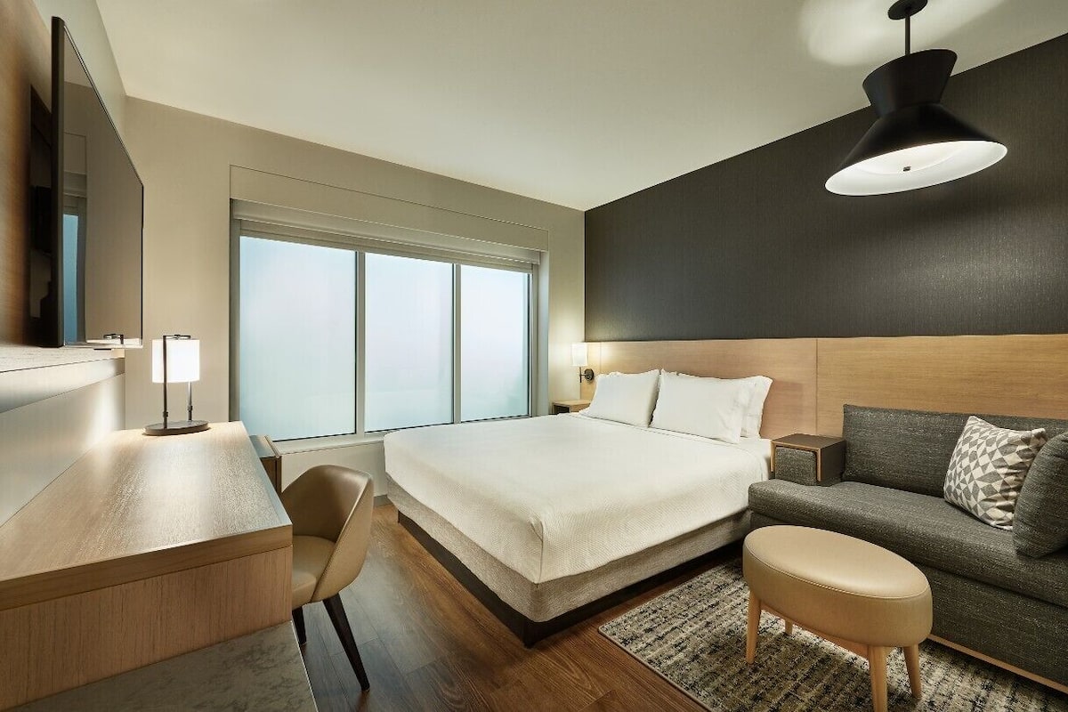 Ett modernt hotellrum med en stor säng, ett skrivbord med en lampa, en soffa och ett stort fönster som släpper in naturligt ljus.
