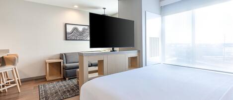 Quarto de hotel com cama, escrivaninha com TV de tela plana, cadeira e uma grande janela com vista para a cidade.