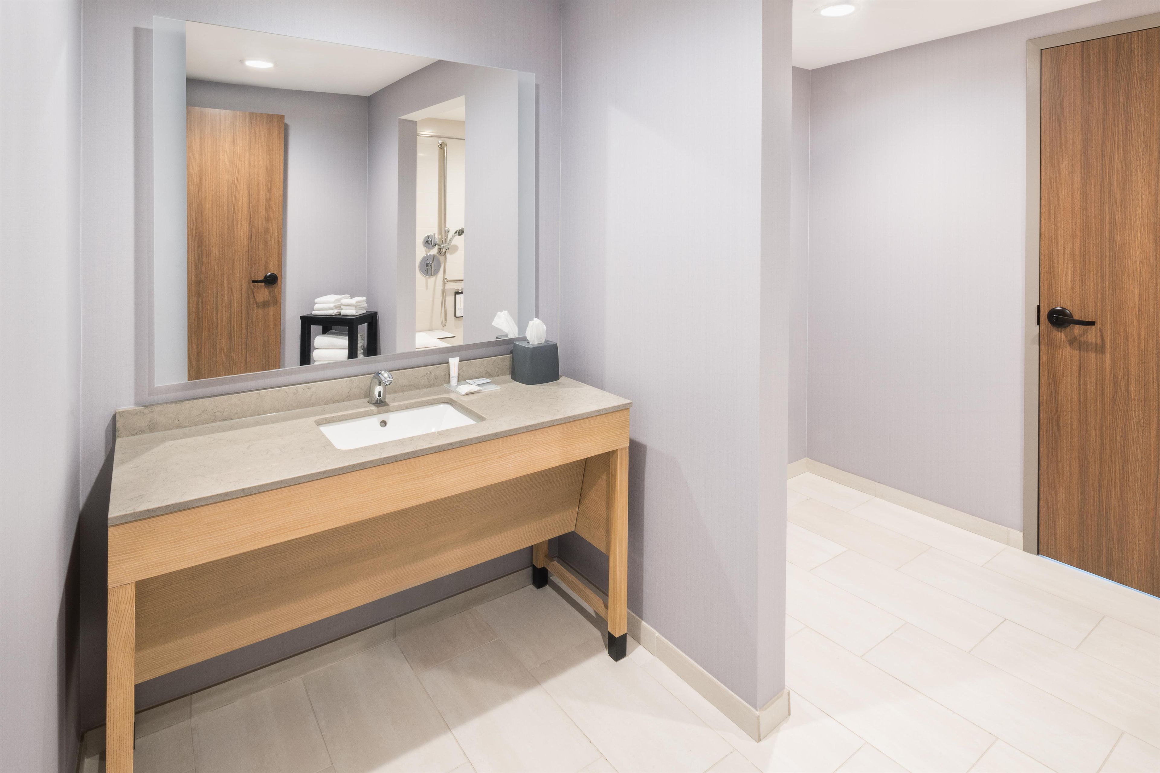 A modern bathroom with a wooden vanity, a large mirror, and a shower visible in the reflection.