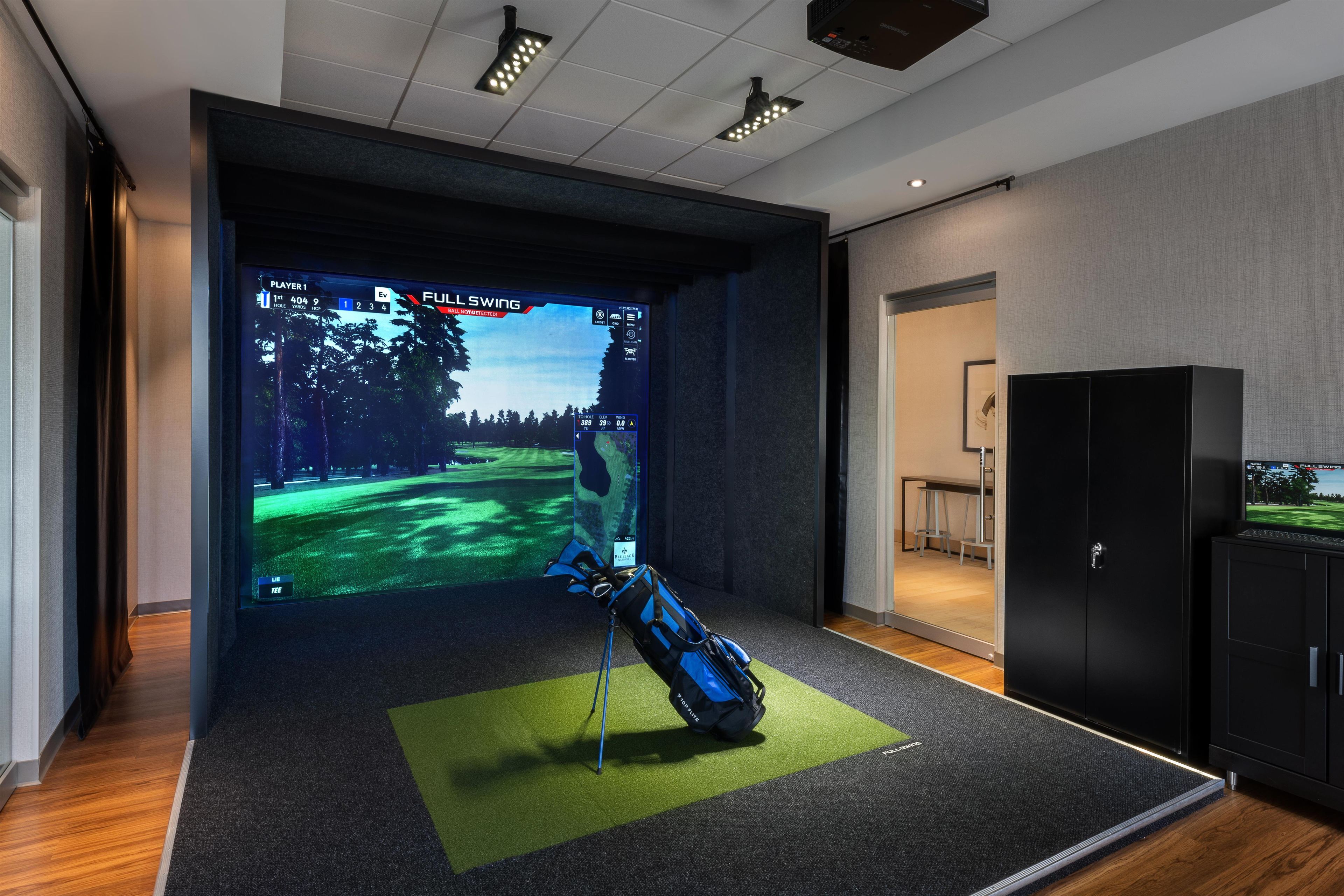 A room with a large screen displaying a golf game, a golf bag, and a cabinet.