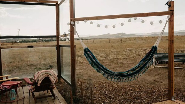 Una hamaca colgada en una estructura de madera, con vista a un campo cubierto de hierba y montañas.