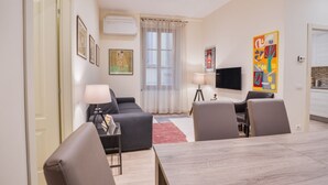 A modern living room with a sofa, a dining table, and chairs. There is a TV, a lamp, and artwork on the walls.