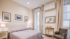 A bedroom with a bed, bedside lamps, a chair, a small table with decorative items, and framed pictures on the wall.