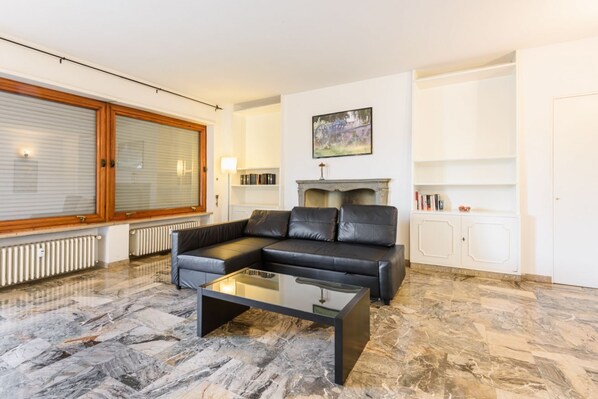 A modern living room with a sectional sofa, a glass coffee table, a fireplace, and a large window with blinds.