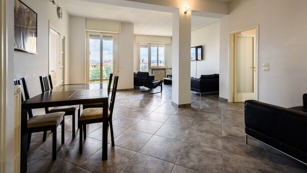 Un salón amplio con una mesa de comedor, sillas, un sofá negro y un televisor montado en la pared.