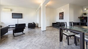 Una sala de estar moderna con una televisión de pantalla plana, un sofá, una mesa de comedor con sillas y un ventanal grande.