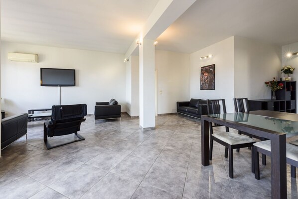 A modern living room with a flat-screen TV, a sofa, a dining table with chairs, and a large window.
