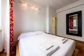 A bedroom with a bed, white linens, a mirror, and a red curtain.