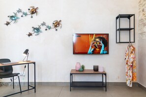 A modern living room with a flat-screen TV, a desk with a chair, a small table with decorative items, and wall-mounted shelves.