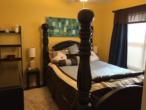 Classic four-poster bed with ornate headboard and animal-shaped pillow, accompanied by a bedside table and geometric-patterned artwork.