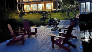 Eine Holzterrasse mit Adirondack-Stühlen und einem Esstisch, mit Blick auf einen See und ein großes Haus mit zahlreichen Fenstern und einer Veranda.