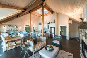 A spacious loft-style living area with a high ceiling, exposed wooden beams, a dining area with a wooden table and chairs, a striped sofa, and a stone fireplace. - Beach House (Huntsville)