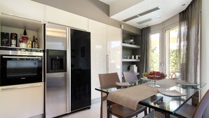A modern kitchen with a built-in oven, refrigerator, and glass dining table with chairs. - Contemporary luxury villa with eclectic design a few steps away from Varkiza bay (Vari)