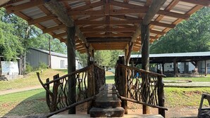 Un pabellón rústico de madera con techo de metal, rodeado de escalones de piedra y baranda de madera.