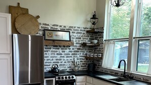 A kitchen with a brick wall, stainless steel appliances, a wooden table, wicker chairs, and a window with a view of greenery.