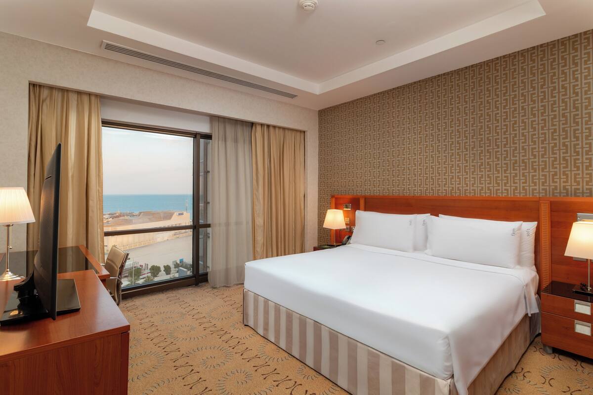 A hotel room with a large bed, a desk with a computer, a TV, and a view of the ocean through the window.