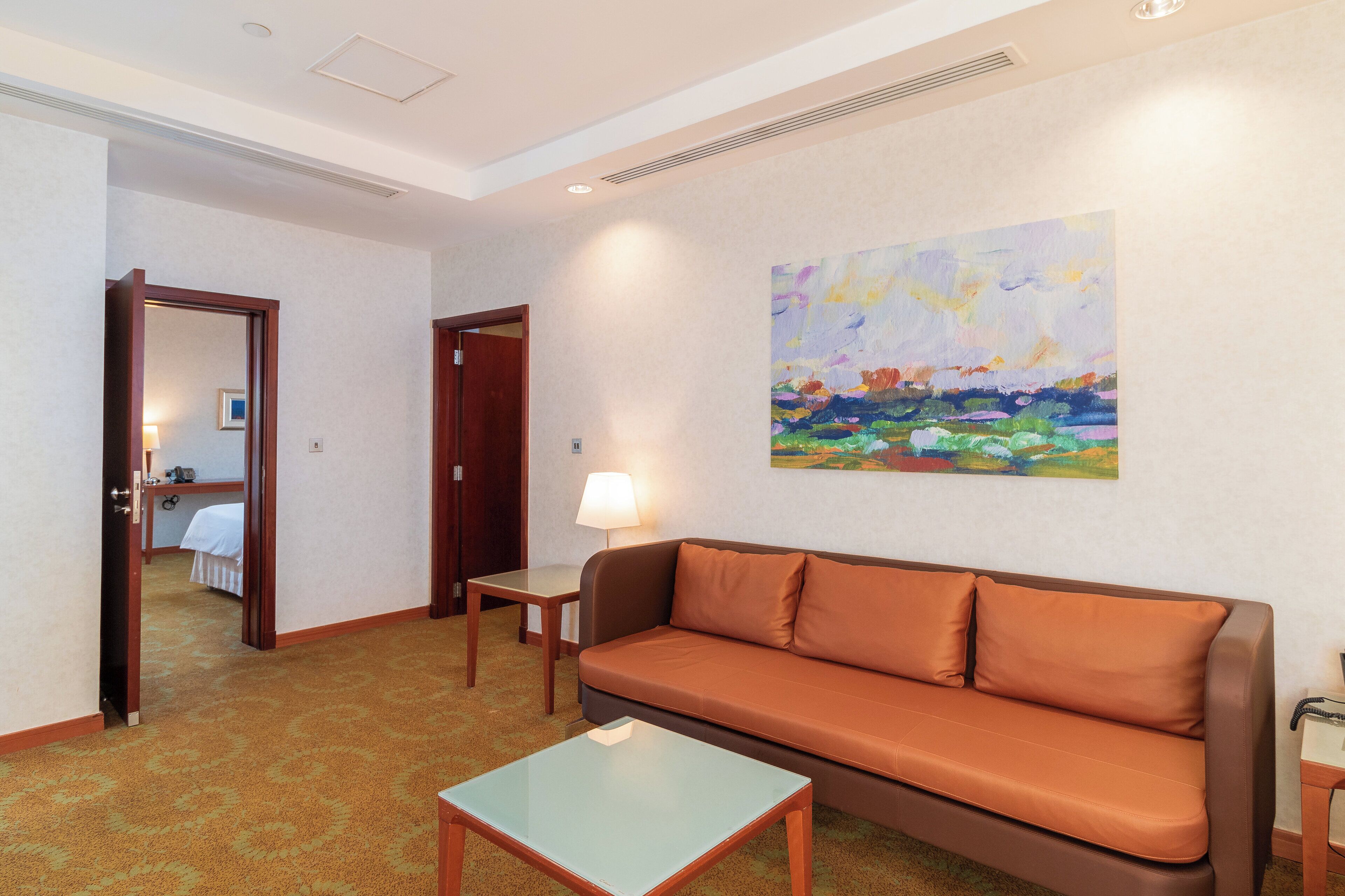 A hotel room with a brown sofa, a glass coffee table, a lamp, and a painting on the wall.