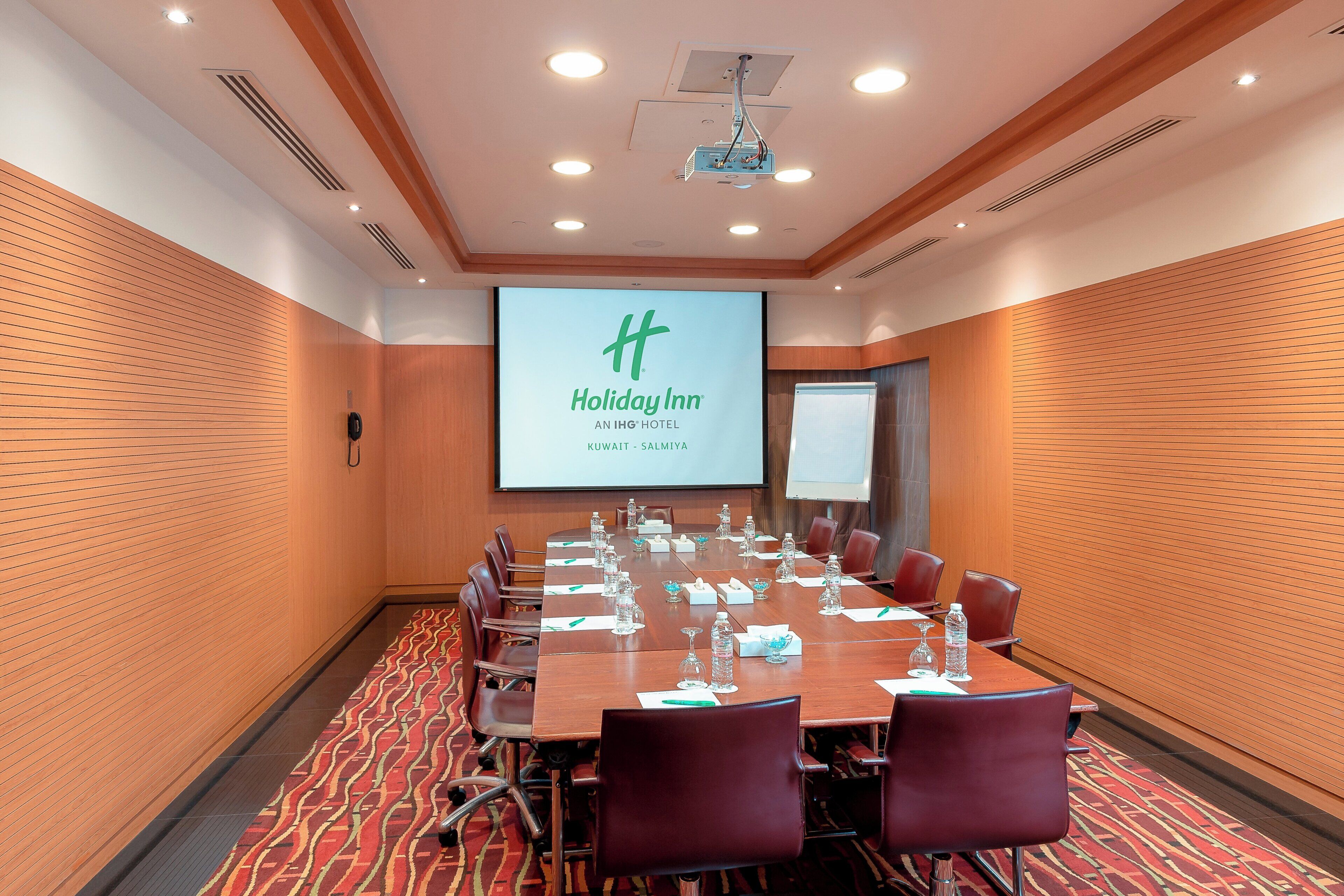 Ein Konferenzraum mit großem Tisch, Stühlen, einer Projektionsleinwand und einem Präsentationsbildschirm.