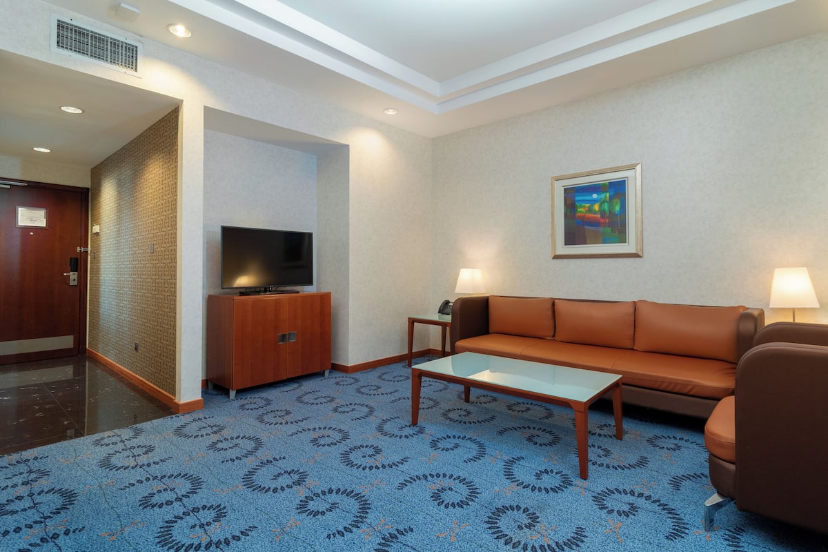 A hotel room with a blue patterned carpet, a brown leather sofa, a glass coffee table, a TV on a wooden cabinet, and two wall lamps.