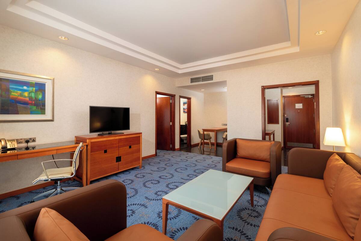 A hotel room with a sofa, armchair, TV, desk, and a patterned carpet.