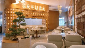 Un restaurante con una pared de madera y el nombre 'SAKURA' iluminado. Se observan mesas preparadas para comer, sillas y un bonsái en primer plano.
