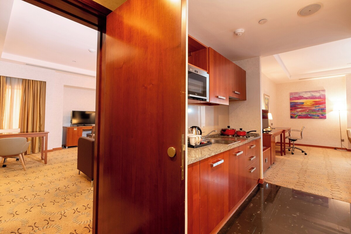 A modern kitchen with wooden cabinets, a microwave, and a sink. There is a dining area with a table and chairs, and a painting on the wall.