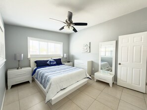 A bedroom with a bed, two bedside tables, a ceiling fan, a window with blinds, a door, and a small bedside table with a lamp.