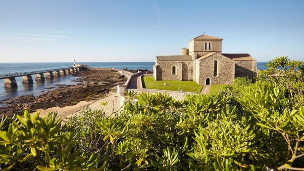 Una iglesia costera con fachada de piedra, un sendero pavimentado que lleva al mar y un césped en primer plano.