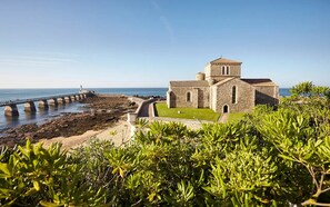 Una iglesia costera con fachada de piedra, un sendero pavimentado que lleva al mar y un césped en primer plano.