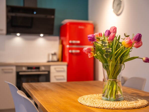 Una cocina con un refrigerador rojo, una mesa de madera con un protector de mesa tejido, y un florero de vidrio con tulipanes sobre la mesa.