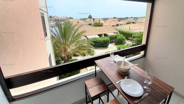 A dining area with a table set for two, a window with a view of a residential neighborhood, and a palm tree outside. - Studio Port Venus Sensation (LE CAP D'AGDE)