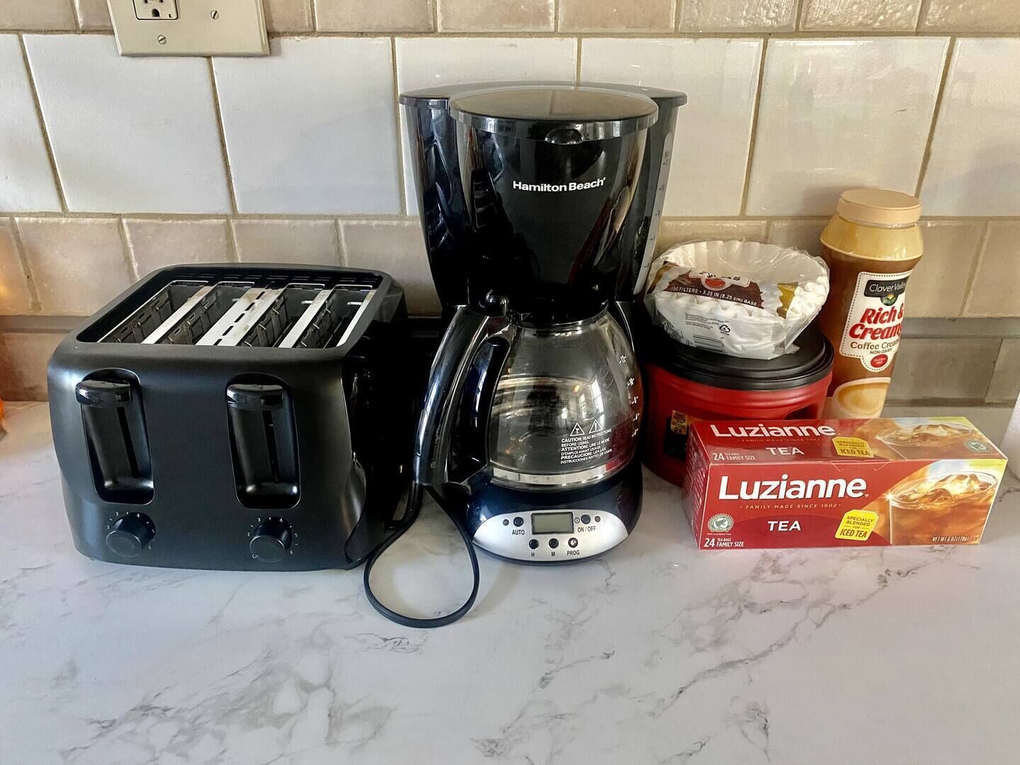 A kitchen with a toaster, a Hamilton Beach coffee maker, a jar of coffee creamer, a box of Lutzianne iced tea, and a container of sugar.