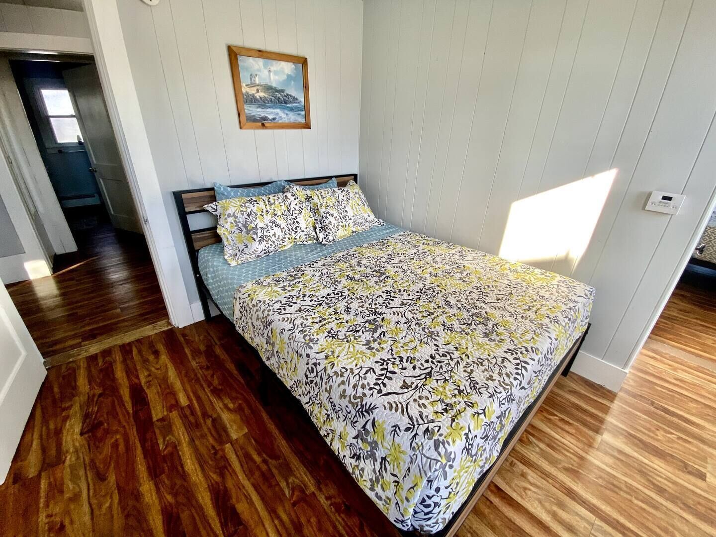 A bedroom with a bed, wooden flooring, a painting on the wall, and a door leading to another room.