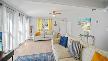 Ein Wohnzimmer mit einer Couch, einem Couchtisch und einem Teppich. Eine Tür ist mit einem 'EXIT'-Schild gekennzeichnet, es gibt einen Deckenventilator und ein Fenster mit Vorhängen.