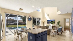 Un'ampia zona giorno con isola cucina, tavolo da pranzo e sedie. Si intravede una piscina attraverso delle grandi porte scorrevoli, e un televisore montato a parete.