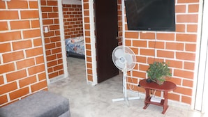 A room with a brick wall, a flat-screen TV mounted on it, a standing fan, a small table with a plant, and a clock on the wall. - Casa Hotel ivanna (Montenegro)