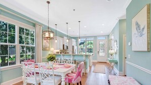 A spacious kitchen with white cabinetry, a dining area with a pink and white table, and a view of the outdoors through large windows.