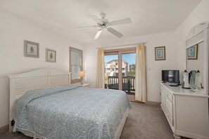 Un dormitorio con cama, ventilador de techo, cómoda, televisor y balcón con vistas a edificios.