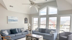 A spacious living room with a grey sofa set, a wooden coffee table, and a wicker chair. - Seven Sisters 26: Oceanside, hot tub, community pool, easy beach access. (Nags Head)