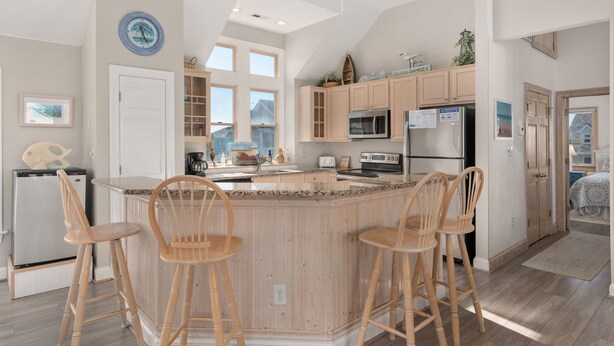 A kitchen with a central island, wooden bar stools, a refrigerator, a microwave, and a clock on the wall. - Seven Sisters 26: Oceanside, hot tub, community pool, easy beach access. (Nags Head)