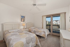A bedroom with two beds, a ceiling fan, a nightstand with a lamp, and a window with a view of houses. - Seven Sisters 26: Oceanside, hot tub, community pool, easy beach access. (Nags Head)