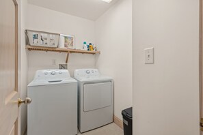 Uma lavanderia com duas máquinas de lavar e secar frontais brancas, uma prateleira com produtos de limpeza e uma lixeira.