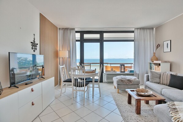 A modern living room with a TV, dining table, and sofa, overlooking a balcony with a view of the sea.