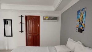Un dormitorio con cama, puerta de madera, aire acondicionado de pared, un cuadro enmarcado y un espejo.