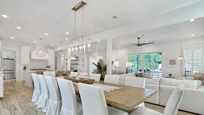 A spacious kitchen with white cabinetry and a central island, a dining area with a wooden table and white chairs, and a living area with a sofa and a ceiling fan.