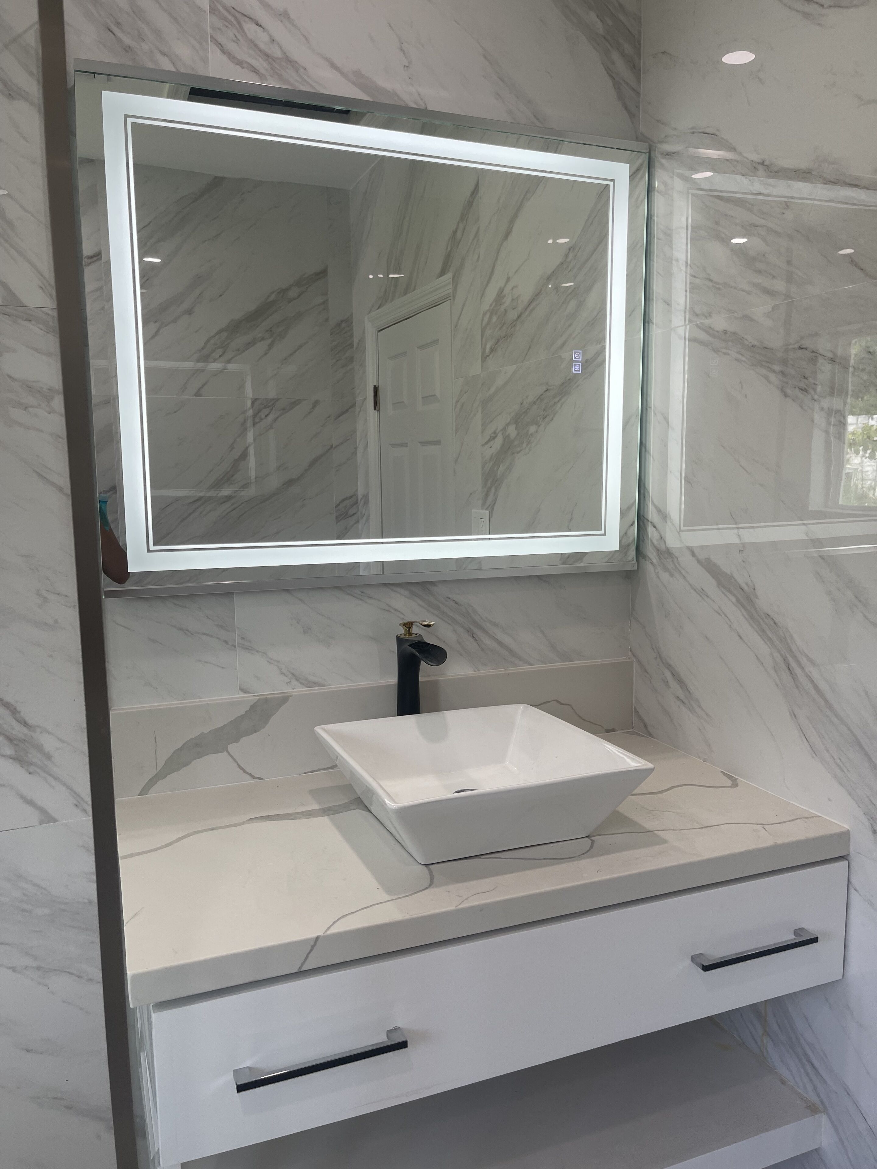 Un baño con lavamanos de mármol blanco, un espejo grande con iluminación integrada y una puerta al fondo.