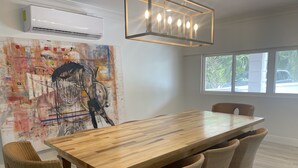 Un comedor con mesa de madera y sillas de mimbre, un cuadro abstracto grande en la pared, y una ventana con vistas al exterior.